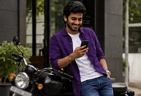 A smiling man in a purple shirt checks his phone, leaning against a motorcycle.