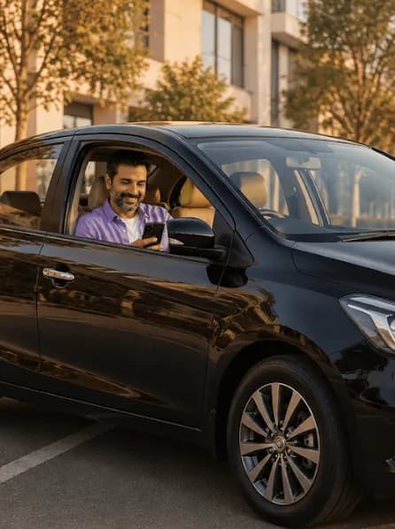 A smiling bearded man in a purple shirt sits in a black car, holding his phone.