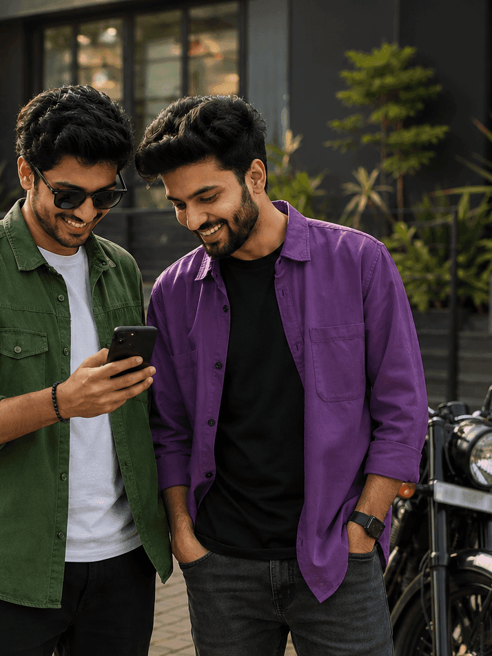 Two young men in casual shirts smiling while looking at a smartphone screen outdoors.